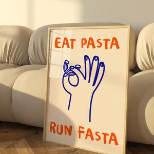 Peut inclure: Une impression encadrée avec un fond blanc et une illustration bleue d'une main tenant un morceau de pâtes. Le texte "EAT PASTA RUN FASTA" est écrit en lettres orange.