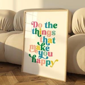 May include: A framed print with a colourful, retro-style font that reads "Do the things that make you happy". The print is leaning against a beige sofa in a room with a wooden floor.