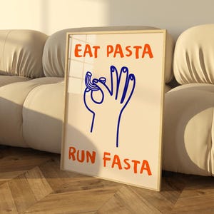 Può includere: Una stampa incorniciata con uno sfondo bianco e un'illustrazione blu di una mano che tiene un pezzo di pasta. Il testo "EAT PASTA RUN FASTA" è scritto in lettere arancioni.