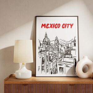 May include: Black and white line drawing of the Mexico City skyline with the text "MEXICO CITY" in red letters. The drawing is framed and hanging on a wall above a wooden shelf with a white lamp and a decorative vase.
