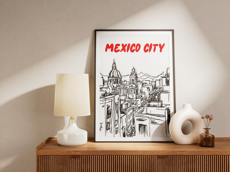 May include: Black and white line drawing of the Mexico City skyline with the text "MEXICO CITY" in red letters. The drawing is framed and hanging on a wall above a wooden shelf with a white lamp and a decorative vase.