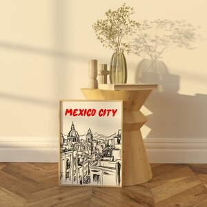 May include: A black and white line drawing of a cityscape with the text "MEXICO CITY" in red letters. The drawing is framed and displayed on a wooden table with a vase of dried flowers.