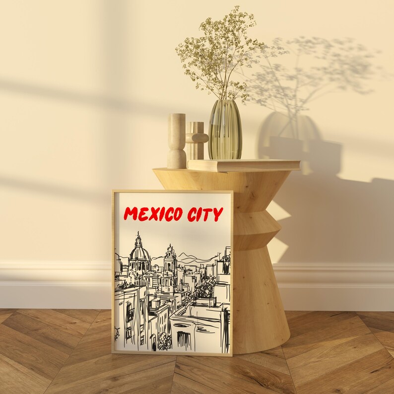 May include: A black and white line drawing of a cityscape with the text "MEXICO CITY" in red letters. The drawing is framed and displayed on a wooden table with a vase of dried flowers.