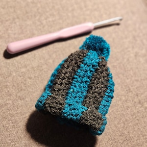 May include: A miniature crochet hat with alternating stripes of teal and dark brown, topped with a teal pom-pom. A pink crochet hook rests in the background, suggesting a handmade craft project.