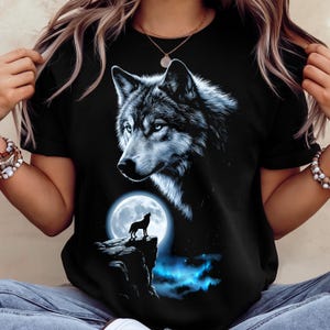 May include: Black t-shirt featuring a detailed illustration of a wolf with blue eyes. The design includes a full moon and a silhouette of a howling wolf on a cliff, with blue and white accents. The shirt is a classic crew neck.