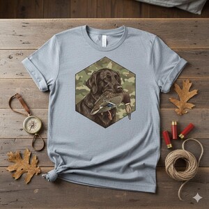 Op de afbeelding: Lichtblauw t-shirt met een afbeelding van een bruine Labrador Retriever die een eend in zijn bek houdt, tegen een camouflage zeshoek. Het shirt is gemaakt van zacht materiaal, ideaal voor dagelijks gebruik.