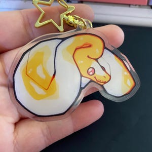 May include: A clear acrylic keychain featuring a cartoon illustration of a white and yellow snake. The snake has a pink eye and a smiling expression. The keychain has a gold star-shaped clasp.