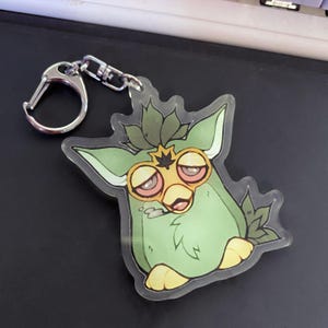 May include: A clear acrylic keychain featuring a green cartoon creature with yellow eyes and a yellow beak. The keychain has a silver metal clasp and chain. The creature has a leaf-like design on its head and is holding a small object.
