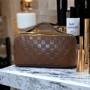 May include: A brown leather travel case with a checkered pattern and gold zipper. The case is sitting on a white marble surface with several bottles of beauty products.