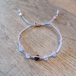 May include: A light blue and pink friendship bracelet with a black lava bead and light blue beads. The bracelet is tied with a brown string and has silver flower charms on each end.