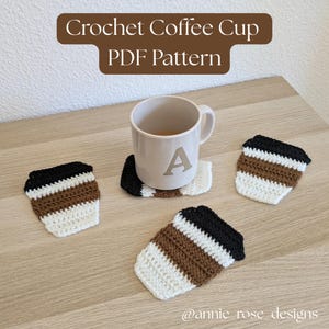 Puede incluir: Una taza de cerámica clara con la letra A impresa en el lateral, rodeada por tres pequeños posavasos de ganchillo con forma de taza de café en negro, marrón y blanco. El texto Crochet Coffee Cup PDF Pattern y @annie_rose_designs es visible.