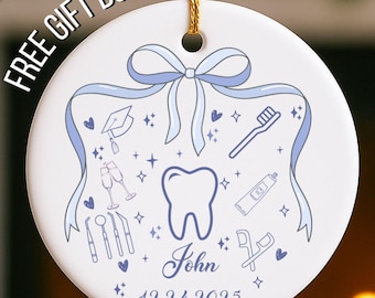 May include: A white ceramic Christmas ornament with a gold string. The ornament features a blue bow, dental tools, and a tooth graphic. The name "John" and the date "12.24.2025" are also displayed.