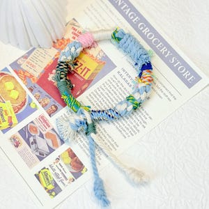May include: A colorful, handmade bracelet featuring a braided design with blue, pink, green, and white yarn. The bracelet is displayed on a vintage-style newspaper with food advertisements.