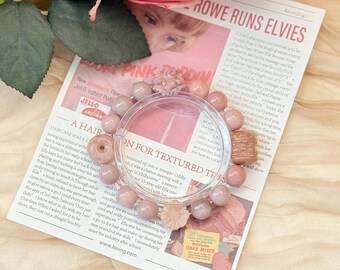 May include: A bracelet with pink beads and decorative charms, including a flower, donut, and a small rectangular shape. The bracelet is displayed on a newspaper with text and a pink rose.