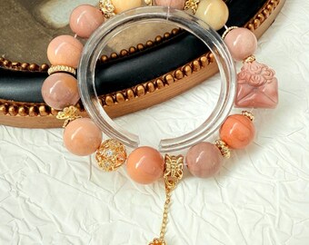 May include: A beaded bracelet featuring a variety of peach, pink, and yellow gemstones. The bracelet includes gold-colored accents and a charm with a flower design. The bracelet is displayed on a white surface.
