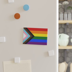 May include: A white refrigerator with a pride flag magnet on the door. The flag is a rainbow with a black and brown triangle on the left side.