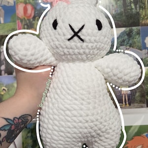 May include: A white crocheted plush rabbit toy with a textured yarn finish, featuring two long ears, a small pink bow on the left ear, and black embroidered eyes and an X-shaped mouth. The text GramsParadise is visible on the side of the plush.