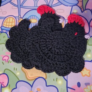 May include: Three black crochet cloud-shaped coasters with red accents on top. The coasters are handmade and have a textured appearance. The background features a colorful, whimsical design with cartoon-like elements.