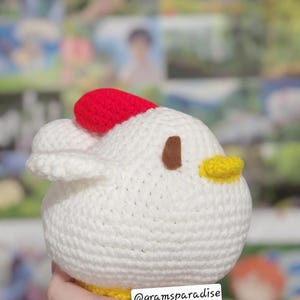 May include: A handmade, crocheted chicken toy. The chicken is white with a red comb, brown eye, and yellow beak. The base of the chicken is yellow. The text "@gramsparadise" is visible on the bottom. The toy is held in a hand.