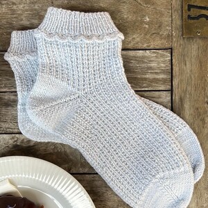 May include: A pair of light gray knitted socks with a scalloped edge, resting on a wooden surface. The socks are next to a white plate with a pastry on it. The socks appear to be handmade.