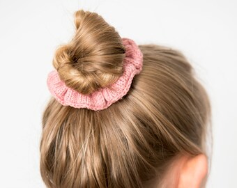 Könnte beinhalten: Eine junge Person mit blonden Haaren, die ein braunes Oberteil und ein rosa gestricktes Haargummi trägt und ihre Haare zu einem Dutt gebunden hat.