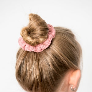 May include: A young person with blonde hair wearing a brown top and a pink knitted hair scrunchie holding their hair in a bun.
