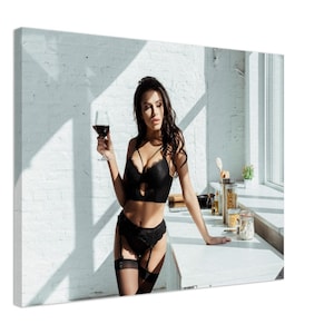 May include: A woman in black lingerie stands in a kitchen, holding a glass of red wine. She is leaning against a white countertop, with a window and white brick wall in the background.