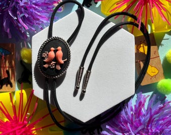 May include: A black bolo tie with a cameo featuring two pink birds kissing. The cameo is set in a silver-toned oval frame.