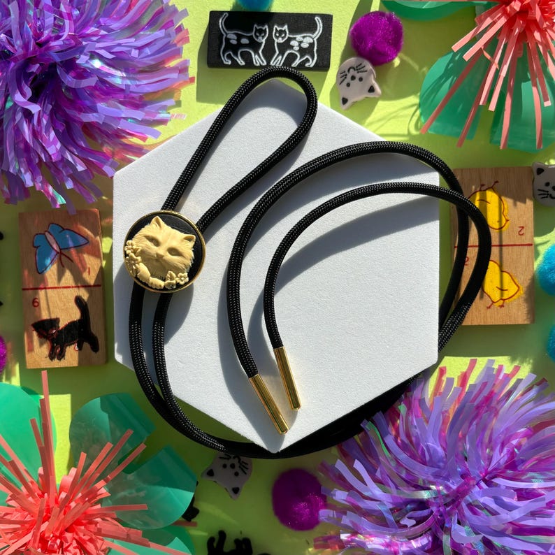 May include: A black bolo tie with a gold-toned cat cameo. The cameo features a white cat with black eyes and a black background. The bolo tie is on a white hexagonal surface.