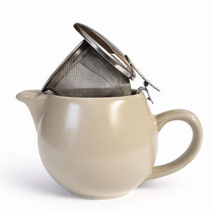 May include: A cream-coloured ceramic teapot with a curved handle and spout. A stainless steel tea infuser with a hinged lid is placed inside the teapot. The infuser is made of fine mesh.