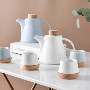 May include: A set of ceramic teapots and cups. The teapots are white and pale blue with wooden lids. The cups are white and pale blue with wooden bases. The set is arranged on a white surface.