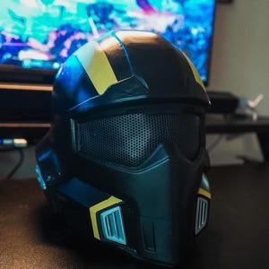May include: A black helmet featuring a matte finish with yellow accent stripes along the top and sides. The front includes a dark mesh visor and a pointed faceplate with two silver-colored vents on the lower sides of the chin area.
