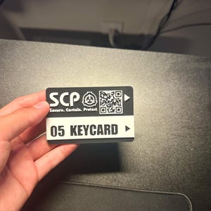 May include: A rectangular black and white plastic keycard featuring the SCP Foundation logo, the text "SCP Secure. Contain. Protect.", a QR code, and the label "05 KEYCARD" printed in bold black lettering on a white horizontal stripe.