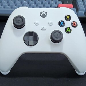 May include: A white wireless video game controller featuring a central Xbox logo button, a directional pad, two black analogue sticks with skull icons, and four coloured action buttons labelled Y, X, B, and A, along with menu, view, and share buttons.