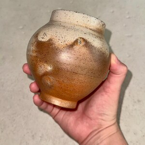 May include: A small, rounded ceramic vase with a narrow neck and a flared rim. The surface features a speckled texture with a color gradient transitioning from a creamy off-white at the top to a warm, earthy brown at the base, showing a mottled finish.