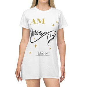 Puede incluir: Vestido camiseta blanco con el texto "I AM Blessed" en oro y negro, junto con un diseño de corazón. El vestido también presenta el texto "John 1:16" y detalles de estrellas doradas.
