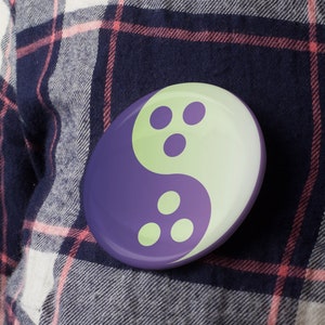 Pode incluir: Um símbolo yin yang roxo e verde com três pontos brancos de cada lado. O símbolo está sobre um fundo de tecido xadrez.
