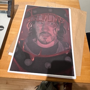 May include: A stack of art prints depicting a man with a beard and dark hair, wearing a dark hat with red accents. The prints are on a wooden table, with a paper cutter and a brown paper envelope nearby.