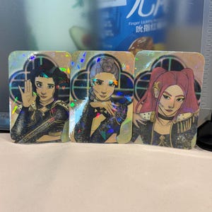 May include: Three holographic trading cards featuring illustrations of women in ornate costumes. Each card has a different woman with unique hairstyles and expressions. The cards have a shimmering, iridescent effect, and are set against a blurred background.