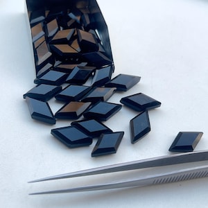Puede incluir: Una colección de pequeñas gemas negras en forma de diamante que se derraman de un recipiente oscuro sobre una superficie blanca. Unas pinzas plateadas descansan cerca, lo que sugiere que se están manipulando las piedras.