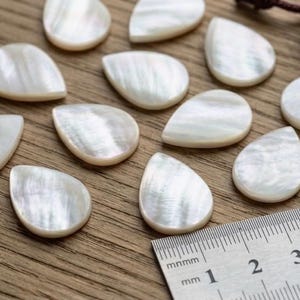 May include: A collection of pearlescent, teardrop-shaped objects, possibly guitar picks, arranged on a wooden surface. A ruler with millimeter markings is visible. A small, dark purple velvet bag is in the background.