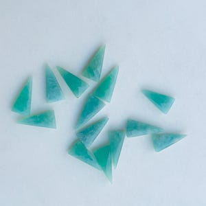 Può includere: Una collezione di pietre preziose triangolari lucide nei toni del verde acqua e turchese. Le pietre sono sparse su una superficie bianca, ognuna con un motivo e una traslucenza unici. Ideali per la creazione di gioielli o l'artigianato.