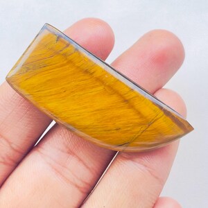 May include: A polished tiger's eye gemstone cabochon with a semi-circular shape, featuring a vibrant golden-yellow hue and characteristic chatoyant bands of dark brown fibers running diagonally across the smooth, reflective surface.
