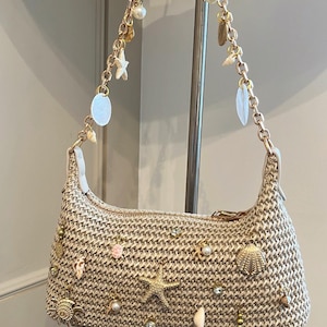 Puede incluir: Un bolso beige tejido con forma curva, adornado con cadenas y abalorios dorados. Los abalorios incluyen conchas marinas, estrellas de mar, perlas y otros elementos decorativos. El bolso tiene una correa blanca y un cierre de cremallera.