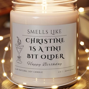 May include: A white soy candle in a clear glass jar with a silver label that reads "Smells Like Christine is a Tini Bit Older Happy Birthday". The candle is 9 ounces and has a burn time of 50 hours.