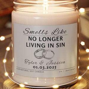 Könnte beinhalten: Eine weiße Sojawachskerze mit einem silbernen Etikett, auf dem "Smells Like No Longer Living In Sin" mit einer Zeichnung von zwei Eheringen steht. Das Etikett enthält auch die Namen "Tyler & Jessica" und das Datum "01.03.2025". Die Kerze ist angezündet und steht auf einer Holzoberfläche.