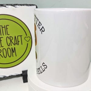 May include: A white ceramic mug featuring a partial view of a graphic with brown and green colors and black text that reads "TTER" and "ELLS" vertically along the side. The mug has a smooth, glossy finish and a cylindrical shape.