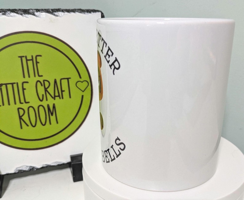 May include: A white ceramic mug featuring a partial view of a graphic with brown and green colors and black text that reads "TTER" and "ELLS" vertically along the side. The mug has a smooth, glossy finish and a cylindrical shape.