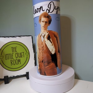 May include: A white tumbler with a blue background and a picture of a man with brown hair wearing a brown vest and a brown jacket. The text "Leon Dyn" is written in black on the tumbler.