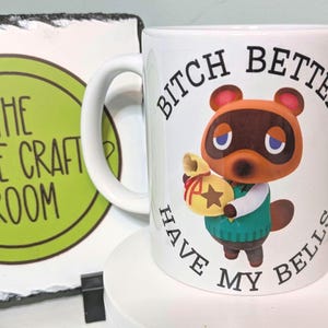 May include: A white ceramic mug featuring a graphic of the character Tom Nook from Animal Crossing holding a yellow bell bag. The text BITCH BETTER HAVE MY BELLS is printed in black capital letters around the character on the front of the mug.
